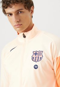 Light orange zip-up jacket with dark blue Barcelona FC logo and Spotify emblem on the chest. Features a high collar and smooth fabric texture.