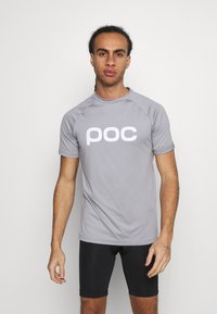 Grå atletisk skjorte med et hvidt "poc" logo, der har korte ærmer og rund halsudskæring. Matchet med sorte tætsiddende shorts.
