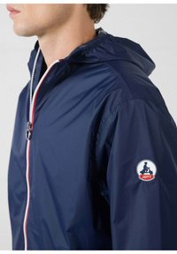 Giacca leggera blu navy con cappuccio, con una cerniera argentata e dettagli rossi. Include una toppa rotonda con il logo sulla manica sinistra.