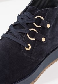 Marinblå mockastövlar med gyllene ögla, ton-i-ton snörning och en kontrasterande blå sula. Har en texturerad yta och rund form.