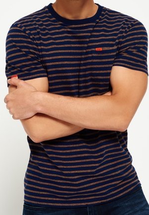 Osoba z założonymi skrzyżowanymi ramionami, ubrana w krótkawy rękaw w granatowo-brązowe paski i ciemnoniebieskie spodnie.