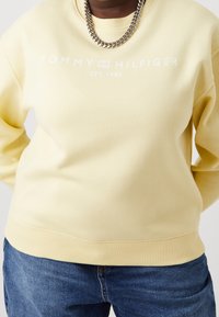 Sweatshirt jaune avec un col et un bas en côtes. Présente le logo blanc "TOMMY HILFIGER" et le texte "EST. 1985". Associé à un jean en denim bleu.