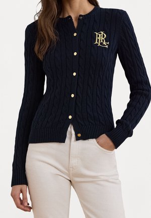 Kvinde iført en sort kabelstrikket cardigan med knapper, guldknapper og logo, kombineret med lyse beige bukser, med hånden i lommen.