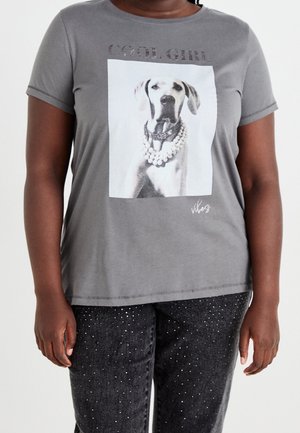 Personne portant un t-shirt gris avec une image d'un chien noir et blanc portant un collier de perles et les mots "COOL GIRL", et un pantalon foncé avec de petites taches brillantes.
