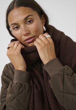 Mujer con pecas que lleva pendientes de aro plateados, múltiples anillos de plata, una chaqueta marrón acolchada y una bufanda gruesa de color marrón oscuro que sostiene cerca de su rostro.