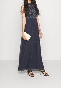 Navy blue maxi dress with a high neckline, embellished beaded bodice, and flowy skirt. Worn with a cream clutch and strappy black heels.