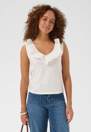Femme aux cheveux bouclés portant un haut blanc sans manches à volants et un jean bleu, tenant un sac à main marron avec un bracelet argenté au poignet.