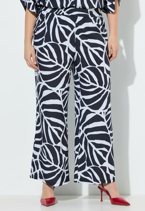 Žena stojící u bílé stěny, oblečená v černobílých kalhotách s širokými nohavicemi a velkým listovým vzorem, s červenými lodičkami na vysokém podpatku s špičatou špičkou.