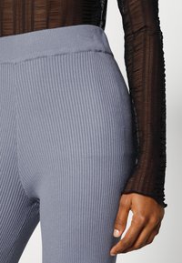 Grå ribbade leggings med bred midjeband har vertikala ränder. En svart genomskinlig långärmad topp med texturerad tyg är delvis synlig.