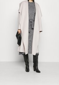 Cappotto lungo, grigio chiaro, con maniche ampie, indossato sopra un vestito grigio scuro con cintura, abbinato a stivali neri fino al ginocchio e una borsa nera.