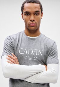 Młody mężczyzna z założonymi rękami, ubrany w szary T-shirt Calvin Klein nałożony na białą koszulę z długim rękawem, na tle białego tła.