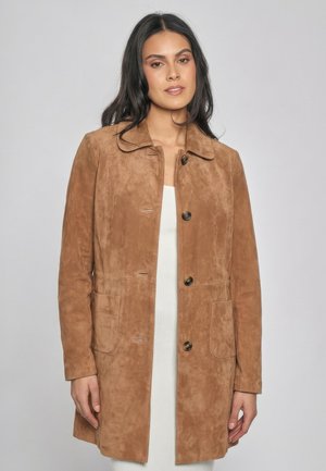 Kvinde med langt mørkt hår iført en brun ruskind frakke med knapper over en hvid ribbet kjole, stående mod en lys grå baggrund.