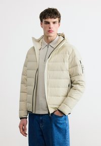 Νεαρός άνδρας που φοράει ελαφρύ μπεζ μπουφάν με επένδυση πάνω από μπεζ πουκάμισο με φερμουάρ και μπλε τζιν, στέκεται μπροστά σε λευκό φόντο.