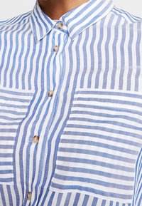 Chemise rayée bleu et blanc avec un col boutonné, deux poches avant, et un tissu en coton texturé aux rayures verticales et horizontales contrastées.