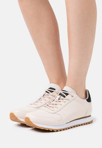 Světle růžové tenisky s texturovaným povrchem, černými akcenty, bílými podešvemi a vzorem vroubkování. Tkaničky jsou ve shodné barvě.