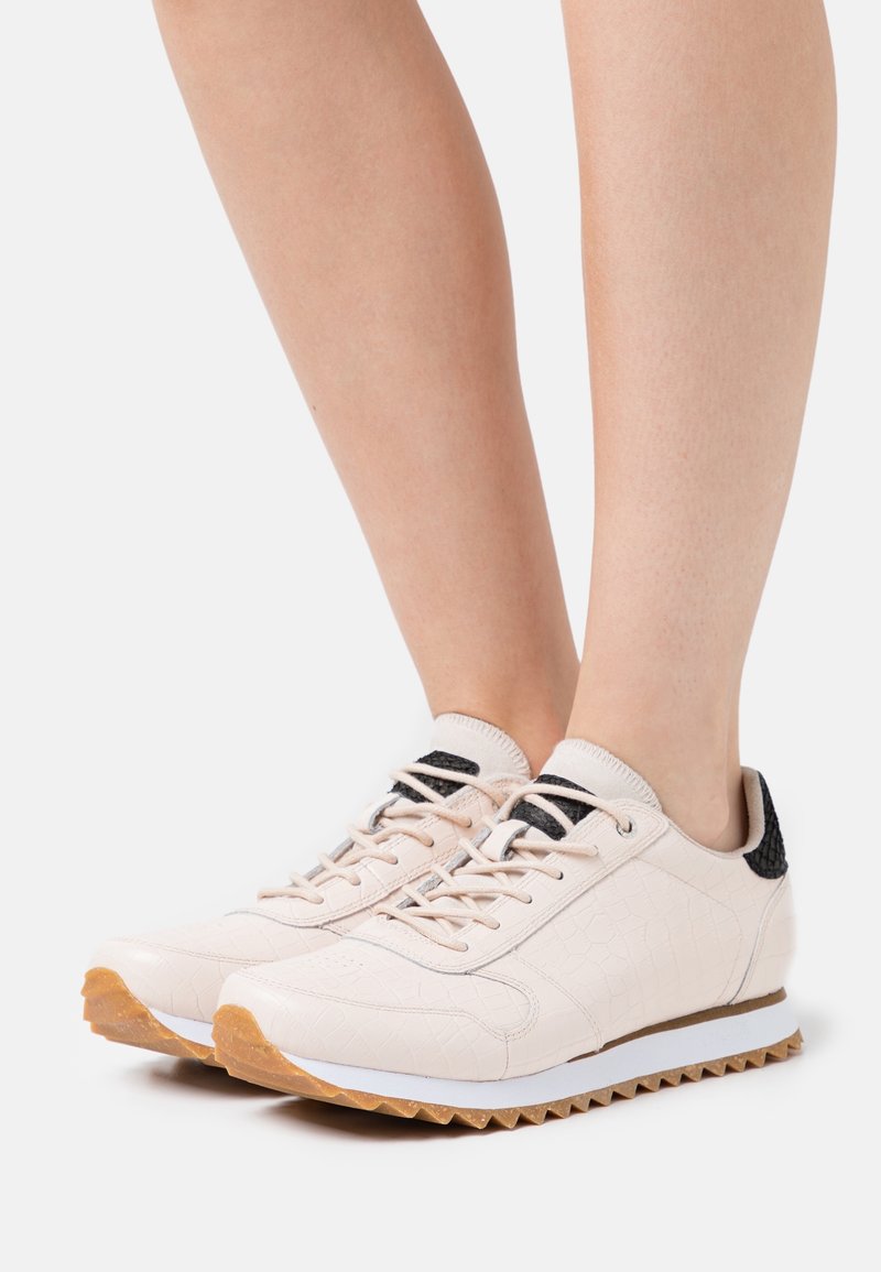 Světle růžové tenisky s texturovaným povrchem, černými akcenty, bílými podešvemi a vzorem vroubkování. Tkaničky jsou ve shodné barvě.