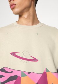 Homme portant un pull beige avec un graphique d'une planète fissurée avec des anneaux roses et des montagnes géométriques colorées en dessous.