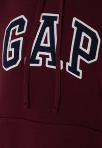 Bordo dukserica s kapuljačom koja sadrži veliki "GAP" logo u bijeloj i tamnoplavoj boji, s prednjim džepom i vezicom za kapuljaču. Tkanina ima mekan, strukturiran završetak.