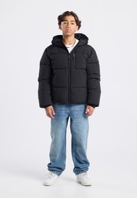 Black padded jacket with a hood, featuring a front zipper and a chest pocket. Worn over a white shirt with blue denim jeans and white sneakers.