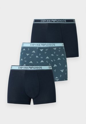 Młody mężczyzna stojący boso, bez koszulki, w niebieskich bokserkach wzorzystych z paskiem "Emporio Armani", patrzący na bok na jednolitym tle.