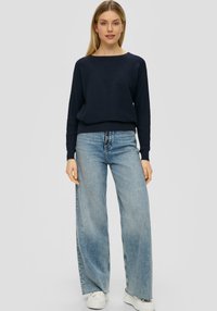 Marineblauer gerippter Pullover mit weitem Halsausschnitt, kombiniert mit hellblauen weit geschnittenen Jeans und weißen Sneakers, in einem lockeren Schnitt.