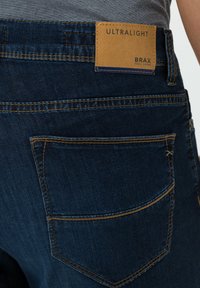 Tummansiniset farkut, joissa on kontrastiväriset keltaiset tikkaukset. Farkuissa on suorakulmainen ruskea nahkaleima, jossa lukee "ULTRALIGHT" ja "BRAX."