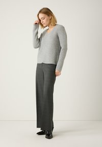 Hellgrauer Strickpullover mit V-Ausschnitt und schrägem Rippenmuster, kombiniert mit hochgeschnittenen strukturierten dunkelgrauen Hosen und spitzen schwarzen Schuhen.