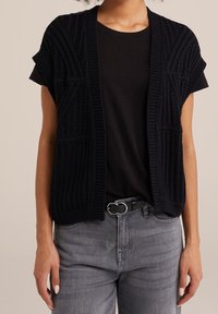 Cárdigan negro de punto con mangas cortas, que presenta patrones texturizados y un frente abierto, llevado sobre una camisa negra y jeans grises.