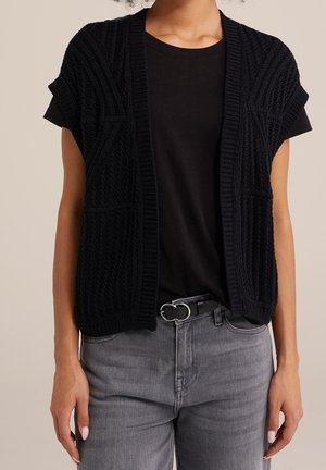 Cárdigan negro de punto con mangas cortas, que presenta patrones texturizados y un frente abierto, llevado sobre una camisa negra y jeans grises.