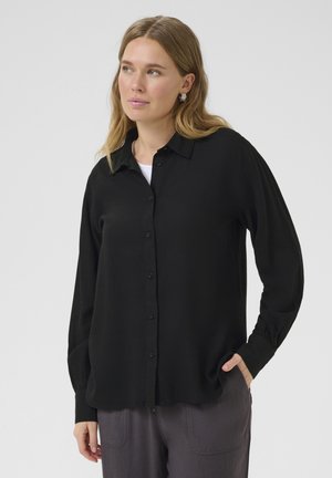 Chemise noire à manches longues, boutonnée, avec une coupe décontractée, comprenant un col, une patte de boutonnage et des détails de poignets froncés. Portée par-dessus un haut blanc.