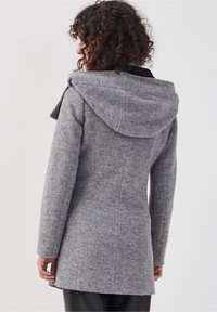 Manteau gris en mélange de laine avec une finition texturée, doté d'une grande capuche et d'une silhouette cintrée, se terminant à mi-cuisse. Détails de couture subtils visibles.