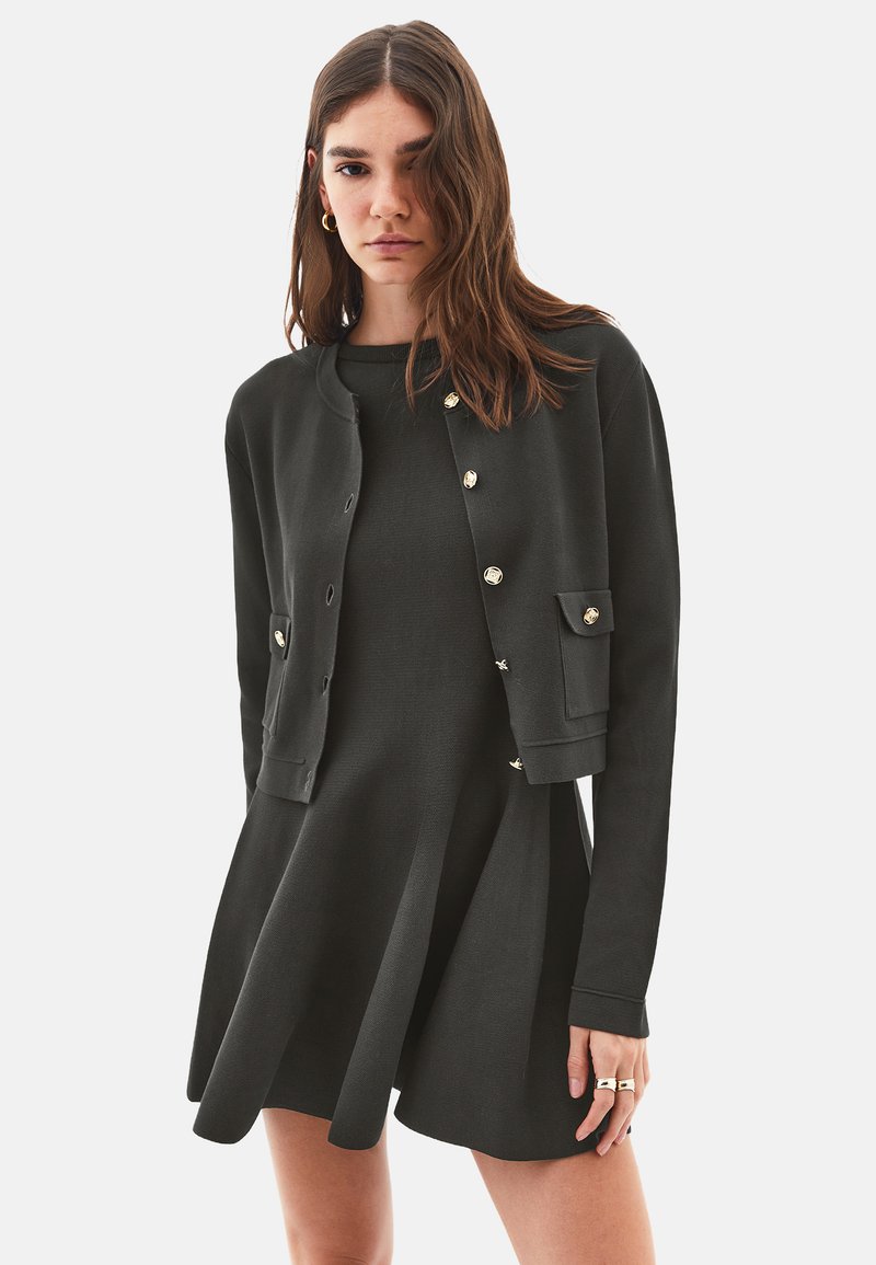 Schwarzes Strickkleid mit tailliertem Oberteil und weit ausgestellt Röcke, kombiniert mit einer kurzen schwarzen Strickjacke mit goldenen Knöpfen und einer Fronttasche.