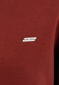 Czerwony bawełniany sweter z małą białą metką z napisem "SMILODOX" i gładką teksturą. Zbliżenie pokazujące detale tkaniny.