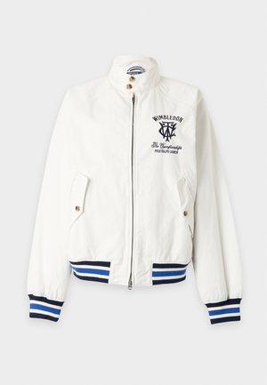 Ung mand iført hvid Wimbledon-jakke, grå skjorte, beige shorts, hvid kasket, hvide sokker og hvide sneakers står med hænderne i lommerne.