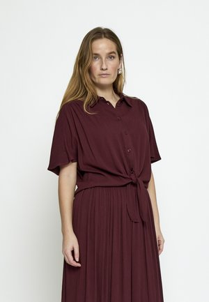 Vrouw met lang lichtbruin haar, gekleed in een bordeauxrood kort mouwtjes hemd met knopen, gestrikt in de taille, en een bijpassende geplooide rok.