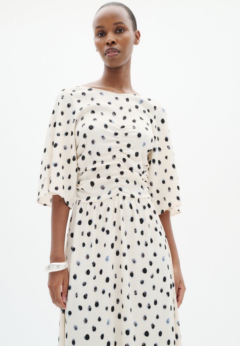 Femme portant une robe blanche à pois noirs et bleu clair, cintrée à la taille, debout avec une expression neutre et un bracelet blanc.