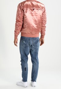 Rosa Satin-Bomberjacke mit geripptem Saum, kombiniert mit hellblauen, abgenutzten Jeans mit Patches und Ausfransungen, ergänzt durch weiße Sneaker.