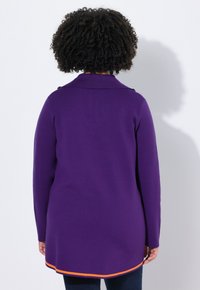 Persoon met krullend haar, gekleed in een lange paarse cardigan met oranje rand, gezien van achteren tegen een plain witte achtergrond.