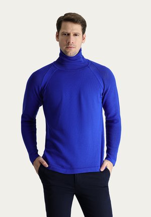 Maglione blu a collo alto con texture a coste e design aderente. Maniche lunghe con cuciture raglan. Abbinato a pantaloni scuri per un contrasto.