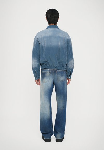 Man standing with back facing camera wearing faded blue denim jacket and loose-fitting jeans with black shoes on white background.