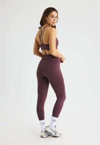 Paarse sportbeha met kruislingse bandjes, gecombineerd met hooggetailleerde paarse leggings. Het model draagt witte sokken en zilveren sportschoenen.