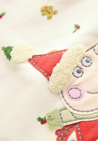 Hvidt stof med et legesygt julesmykke design, der har en rød hat, cremefarvet faux fur kant og farverige broderede detaljer, herunder småkager og kristtorn.