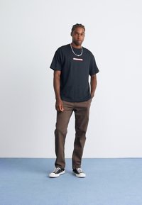 Homme debout sur un sol bleu, portant un t-shirt graphique noir, un pantalon marron, des baskets noir et blanc, avec une chaîne en collier et des cheveux tressés.