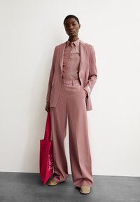 Rosa maßgeschneiderter Anzug mit weit ausgestellten Hosen und einem gestreiften Button-up-Shirt. Das Modell hält eine leuchtend rosa Tote-Bag. Glatte Stoffstruktur.