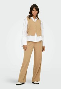 Gilet beige avec cinq boutons sur une chemise blanche, associé à un pantalon large beige et des chaussures blanches. Tissu lisse avec une texture minimale.