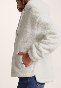 White plush hooded jacket with fleece texture, front pocket, and drawstring detail. Loose fit and rounded hemline.
