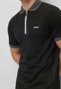 Černé polo tričko s krátkým rukávem, bílým pruhovaným límcem a manžetami, s polovičním zipem a logem "BOSS" na levé hrudi.