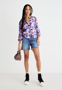 Camicia floreale viola con maniche lunghe, abbinata a pantaloni shorts in denim. La donna tiene in mano una borsa a pochette marrone; indossa sneaker alte nere con suola bianca.