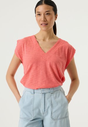 Koraalrode top met V-hals en korte, kapmouwen van gestructureerde stof. Gecombineerd met lichtblauwe, hoogtalgende broek met zijzakken.