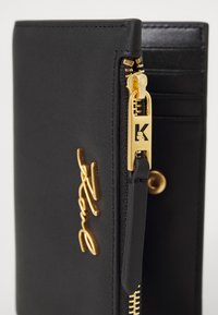 Black leather wallet with gold hardware, featuring a zip closure and a decorative gold script accent. Includes card slots on the interior.